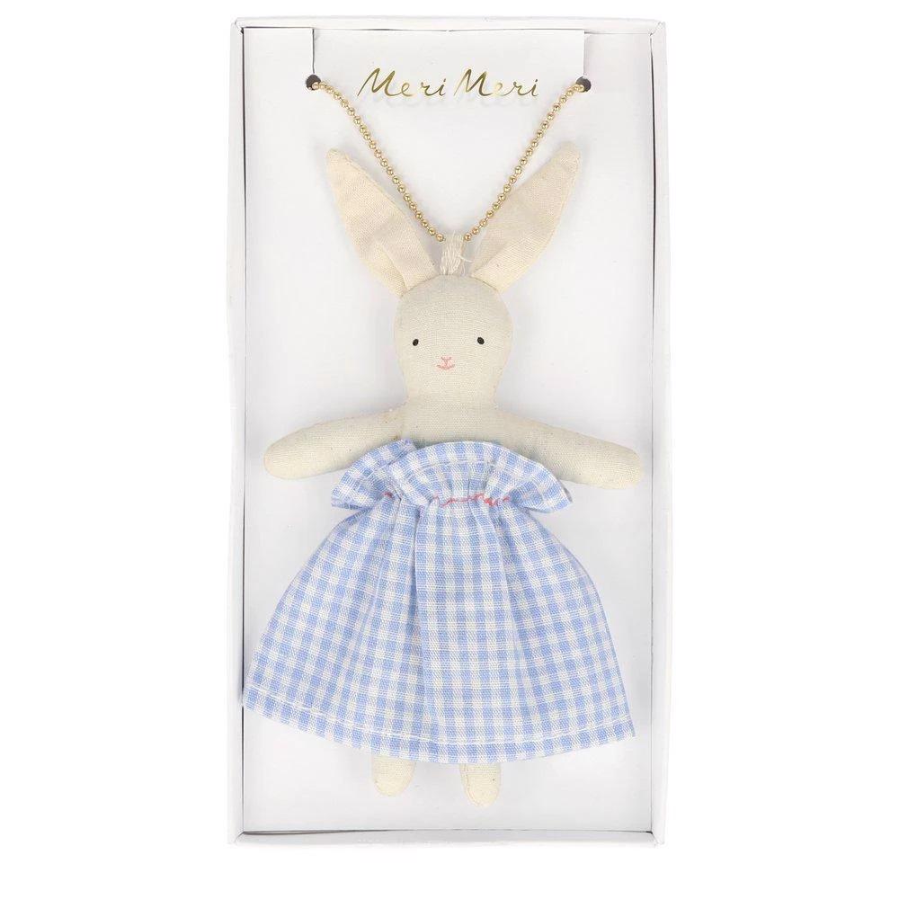 The NECKLACE BUNNY DOLL by MERI MERI is displayed in a rectangular box. The bunny, with long beige ears, is dressed in a blue and white checkered dress. "Meri Meri" is elegantly written in gold lettering above the doll. Perfect as an Easter present, it comes attached with a bead chain.