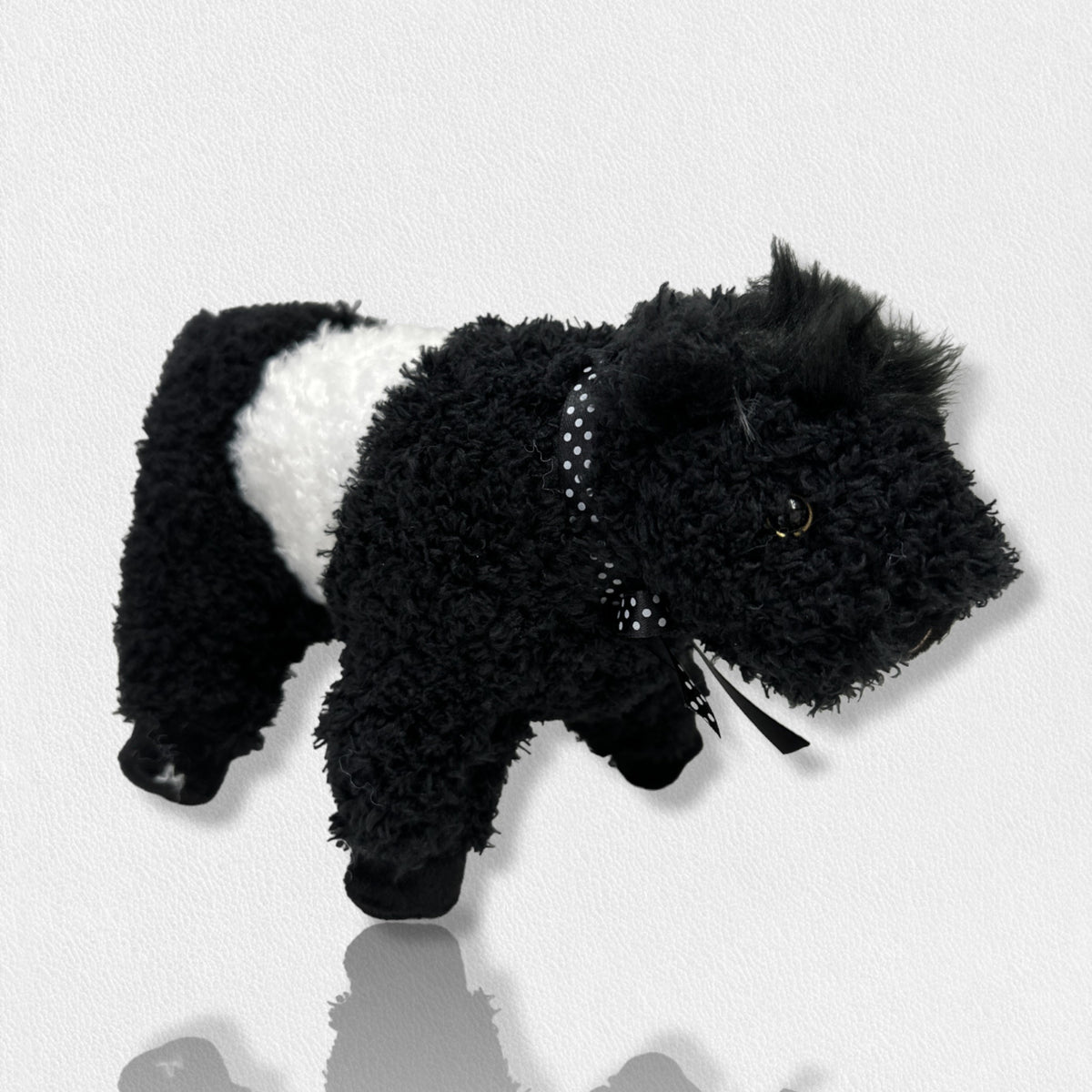 A soft plush toy from the Beverly Hills Teddy Bear Co&#39;s Fearrington Signature Collection, resembling a black and white Belted Galloway cow, stands against a plain white background. This furry mascot features black fuzzy fur on its head, legs, and tail, with a white fuzzy band around its midsection, characteristic of the Galloway breed.