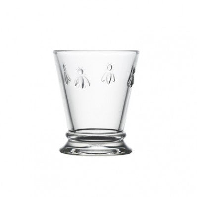 Introducing the BEE MINI TUMBLER by LA ROCHERE: a clear glass tumbler with an outward flare at the top and a thick, round base. This versatile piece of glassware features an embossed pattern of small bees spaced evenly around the upper portion, set against a plain white background.