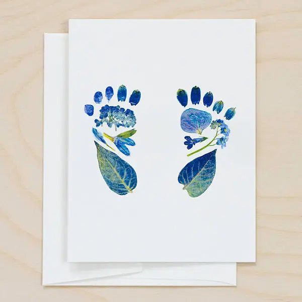The PETAL PEOPLE PRESS - IT'S A BOY CARD from PETAL PEOPLE PRESS showcases two blue baby footprints crafted from pressed flowers and leaves, artfully arranged on a pristine white background. The blank greeting card is displayed on a light wood surface with a matching white envelope situated beneath it.