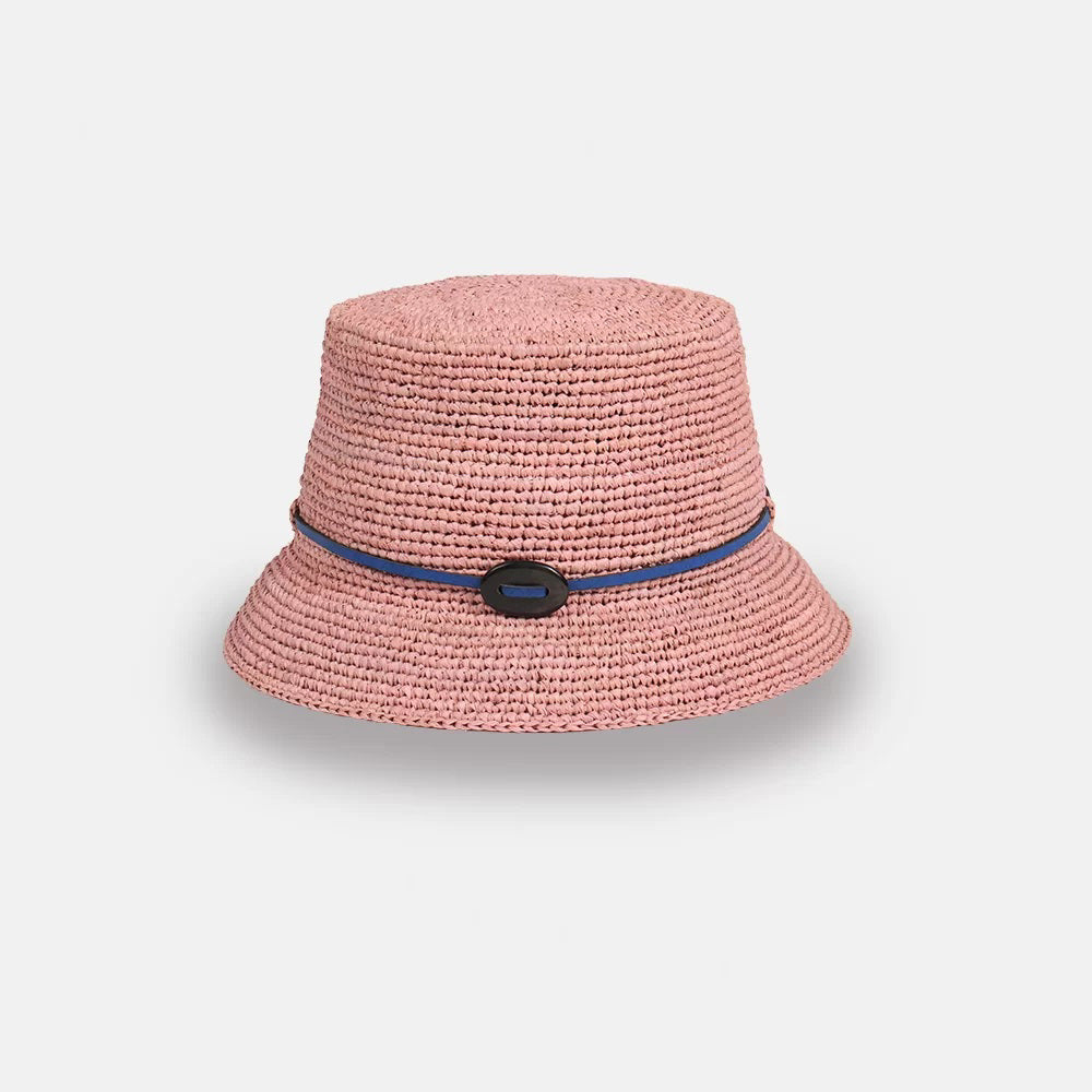 The LEO BUCKET HAT by SANS ARCIDET is a handmade crocheted bucket hat in pink featuring a slightly flared brim. This women's accessory boasts a thin blue cord woven around the base and an oval-shaped black decoration at the front. Crafted from natural raffia, it stands out beautifully against the plain white background.
