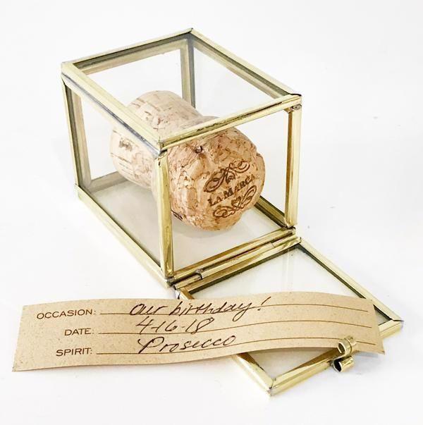 A champagne cork from JM PIERS' LARGE CORK SAFE, enclosed in a small glass display box with golden edges. In front of the box, a brown tag reads: "Occasion: Our Birthday!, Date: 4/6/20, Spirit: Prosecco." It's the perfect gift to commemorate special occasions.