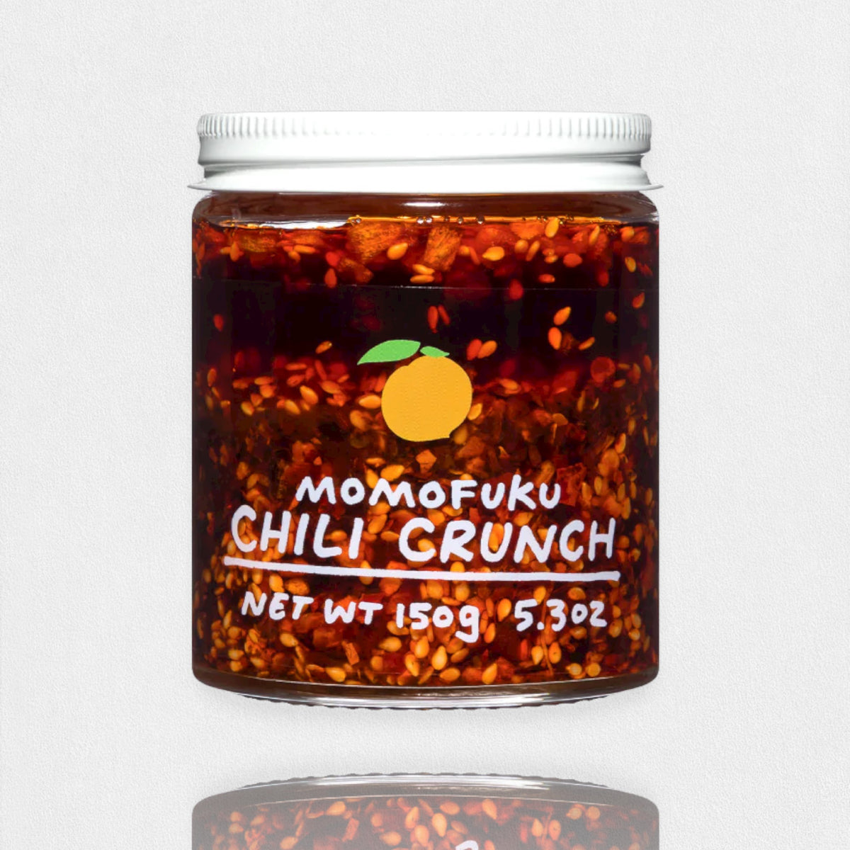 A jar of MOMOFUKU - CHILI CRUNCH, featuring a white lid filled with chili oil and visible chili flakes. The label showcases an orange fruit graphic and text that reads &quot;MOMOFUKU - CHILI CRUNCH, Net Wt 150g (5.3oz).&quot; Made with umami-rich ingredients, it&#39;s perfect for fans of Chinese chili crisp sauces. The background is plain white.