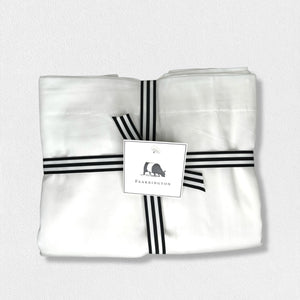 A neatly folded set of white cotton sateen pillowcases from the Fearrington Lifestyle Bedding Collection by Frette North America, boasting a luxurious 300 thread count, is tied with a black-and-white striped ribbon. Attached to the ribbon is a label featuring a small black silhouette of a person seated next to a large bear and the text "Fearrington." The background is light gray.
