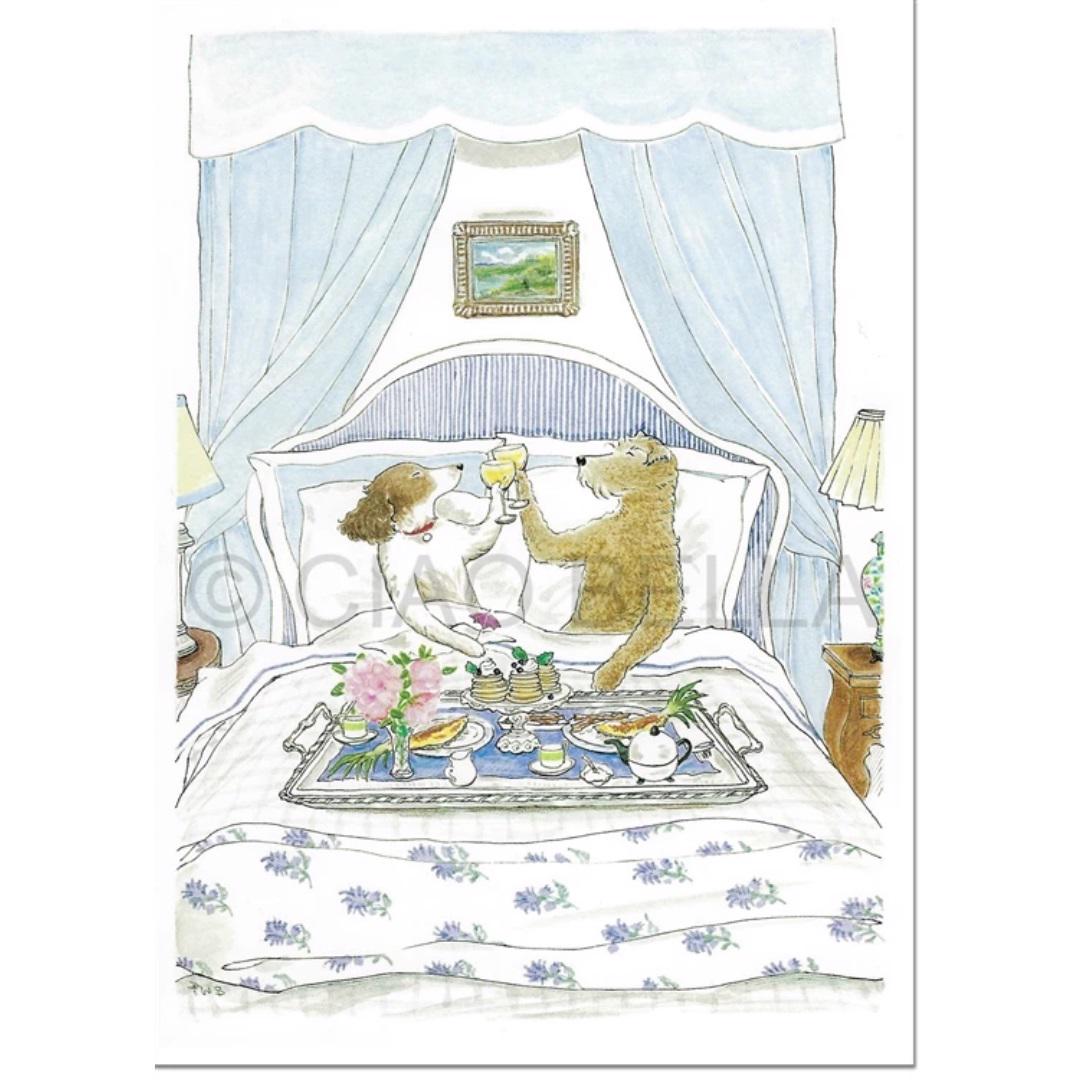 An illustration on the CIAO BELLA - BREAKFAST IN BED CARD by CIAO BELLA shows a dog and a bear sitting up in bed in a cozy bedroom, toasting with drinks. They are surrounded by plates of food on a tray placed over the bed. A blank note card rests nearby on the nightstand with a lamp, and the room features pale blue decor with a picture hanging above the bed.