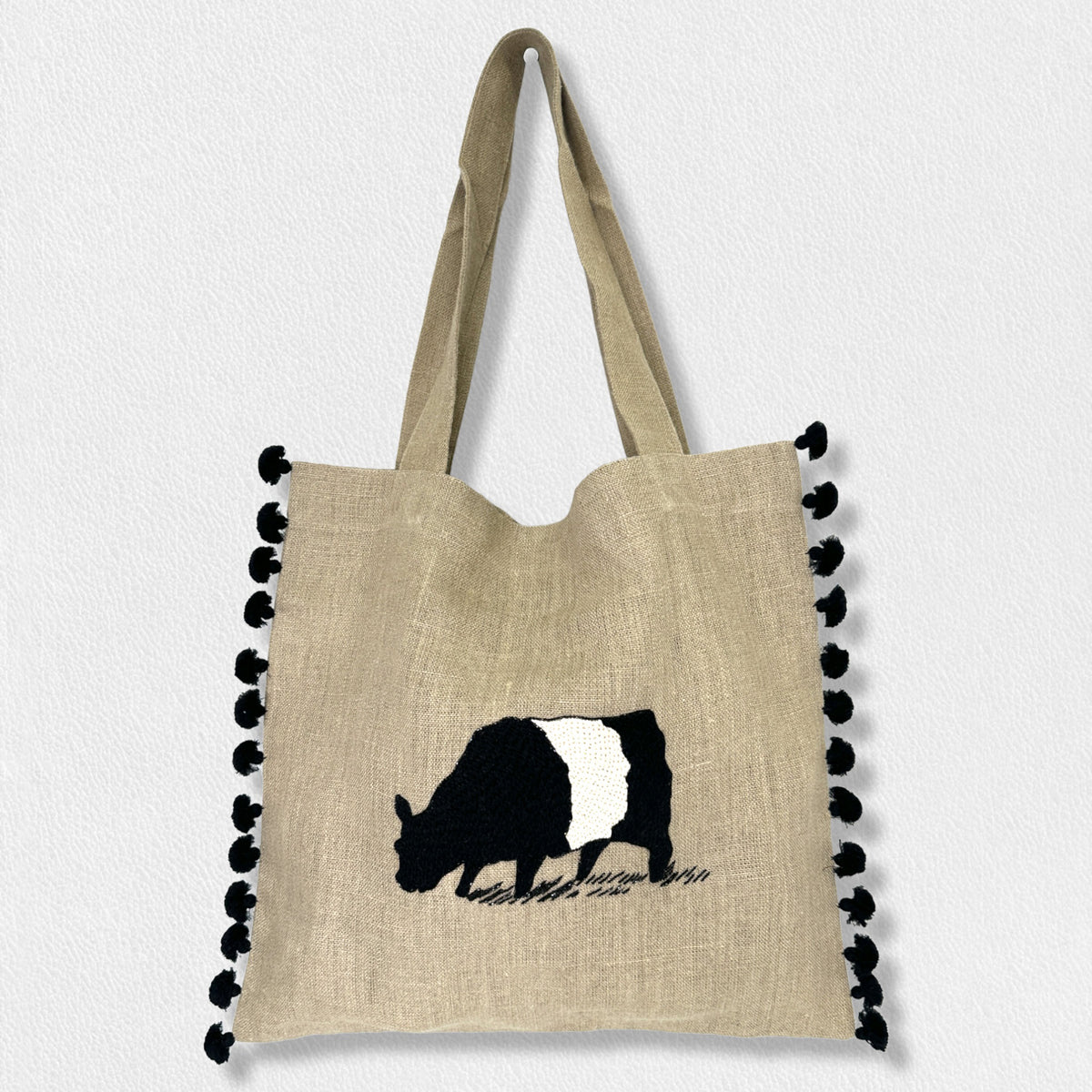 The Signature Beltie Tote from Citrus Fashions&#39; Fearrington Lifestyle Collection is a beige tote bag adorned with black pom-pom trim on the edges and features an embroidered design of a grazing black and white cow. Hand-stitched and perfect for everyday errands, this stylish tote also comes with two handles at the top.