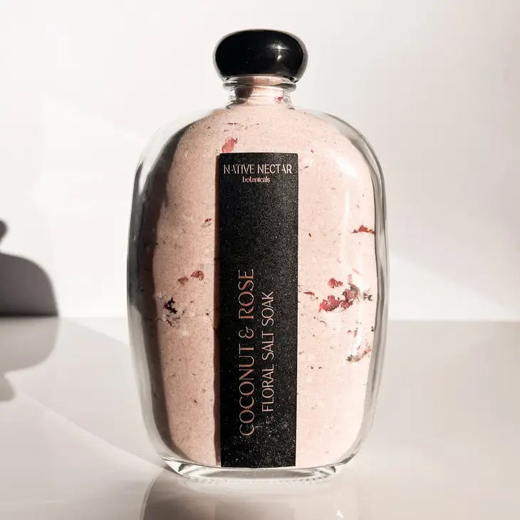 A clear glass bottle contains a pink floral salt soak. The label reads &quot;NATIVE NECTAR,&quot; and in larger font, the product is identified as &quot;COCONUT &amp; ROSE FLORAL SALT SOAK.&quot; Inside are luxurious Himalayan Pink Salt and visible rose petals. The bottle is topped with a black cap and features tinted pink contents.
