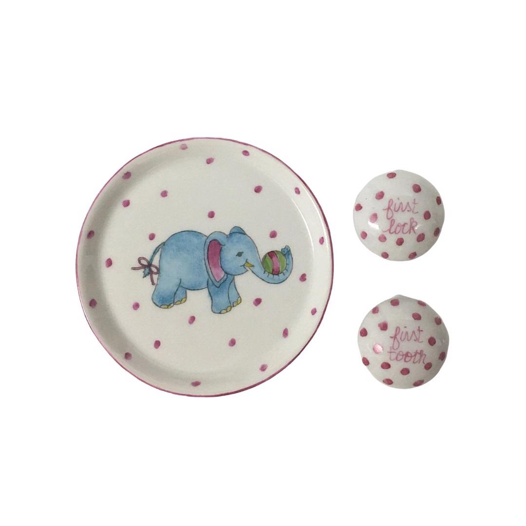 Introducing the TOOTH AND LOCK ELEPHANT (PINK) by ANN MARIE MURRAY, L.L.C., a bespoke porcelain set that includes a beautifully crafted plate and two small lidded containers. The plate showcases a hand-painted blue elephant with pink ears playing with a colorful ball, all surrounded by charming pink polka dots. The dotted lidded containers are thoughtfully labeled "first lock" and "first tooth.