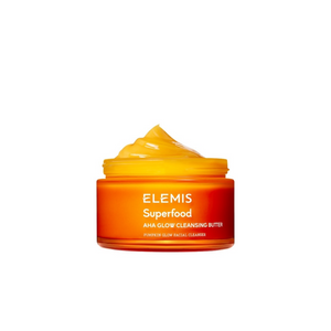 A bright orange jar with a white lid, labeled "ELEMIS - SUPERFOOD AHA GLOW CLEANSING BUTTER" in white text, from the brand ELEMIS - STEINER, featuring nourishing Mango Butter and Pumpkin enzymes. The jar is centered against a plain white background.