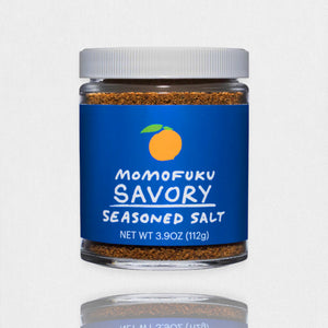 A clear jar with a white lid holds brown MOMOFUKU - SAVORY SALT, featuring a blue label with an orange fruit graphic and "Momofuku Savory Seasoned Salt" text. Net weight: 3.9 oz (112g). Made with premium kosher salt. Brand: MOMOFUKU.