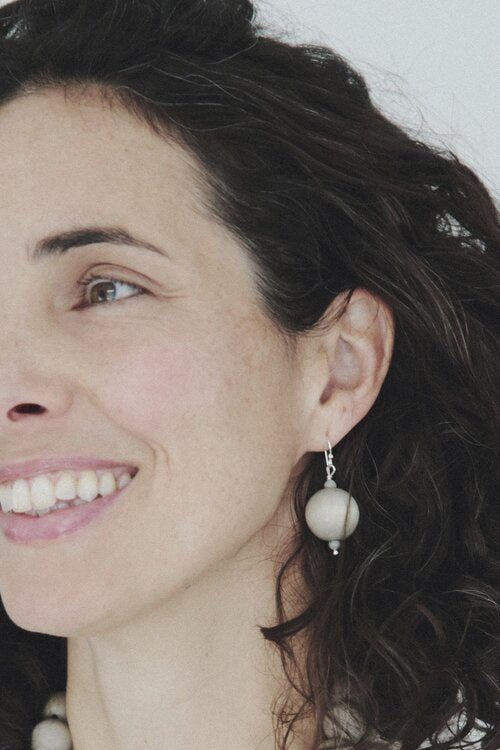 The ANN LIGHTFOOT - GREY WOOD DROP EARRING features large, round light beige beads as the main attraction. Each earring includes two smaller light grey wood beads above and below the central bead. The beads are elegantly attached to a sterling silver earwire, all set against a plain white background.