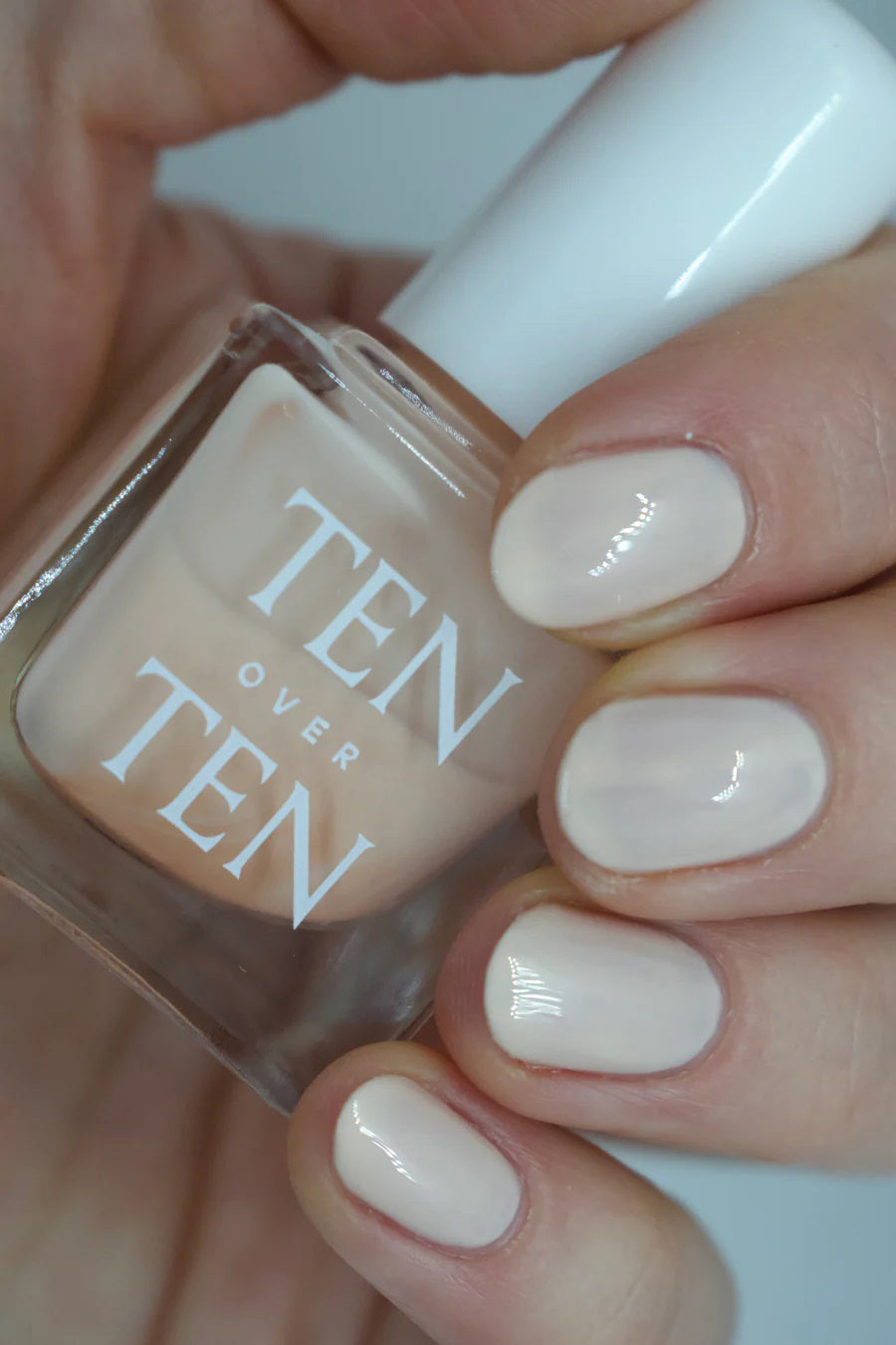 A glass bottle of TENOVERTEN - NAIL POLISH from the brand TEN OVER TEN, featuring a white cap and a dusty pink nail color. The front label displays "TEN OVER TEN" in white uppercase letters. This vegan, eight-free formula ensures both beauty and peace of mind. The background is plain and light-colored.