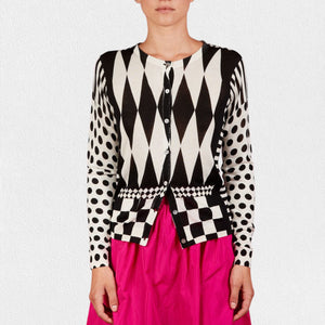 A person wearing the IN BED WITH YOU - GEOMETRIC PRINT CARDIGAN from the Italian brand IN BED WITH YOU/CLAUDIOLAI SRL, featuring alternating diamond shapes on the front, polka dots on the sleeves, and a checkerboard pattern at the hem. They are also wearing a bright pink skirt.
