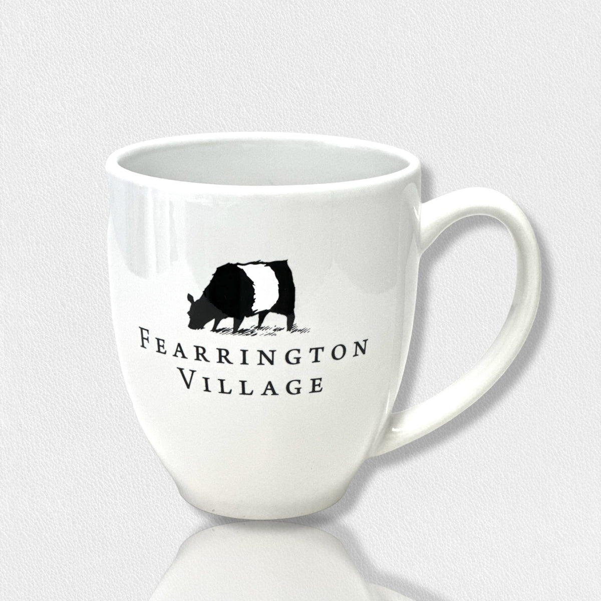 The 16 oz white ceramic coffee mug from the FEARRINGTON SIGNATURE COLLECTION - FEARRINGTON COFFEE MUG 1 COW 3RD EDITION by STRAN PROMOTIONAL SOLUTIONS showcases an illustration of a black and white belted galloway cow, with &quot;Fearrington Village&quot; printed below. This charming mug has a large handle and is displayed on a reflective surface, emphasizing its delightful design.