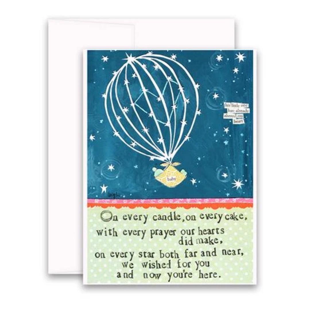 The CURLY GIRL DESIGNS - WISHED FOR YOU CARD by CURLY GIRL DESIGN, INC features a charming nighttime sky background. A heart-shaped hot air balloon carries a small basket with a baby inside, adorned with a tag that reads “baby.” The heartfelt text on the card reads: “On every candle, on every cake, with every prayer our hearts did make, on every star both far and near, we wished for you and now you’re here.”