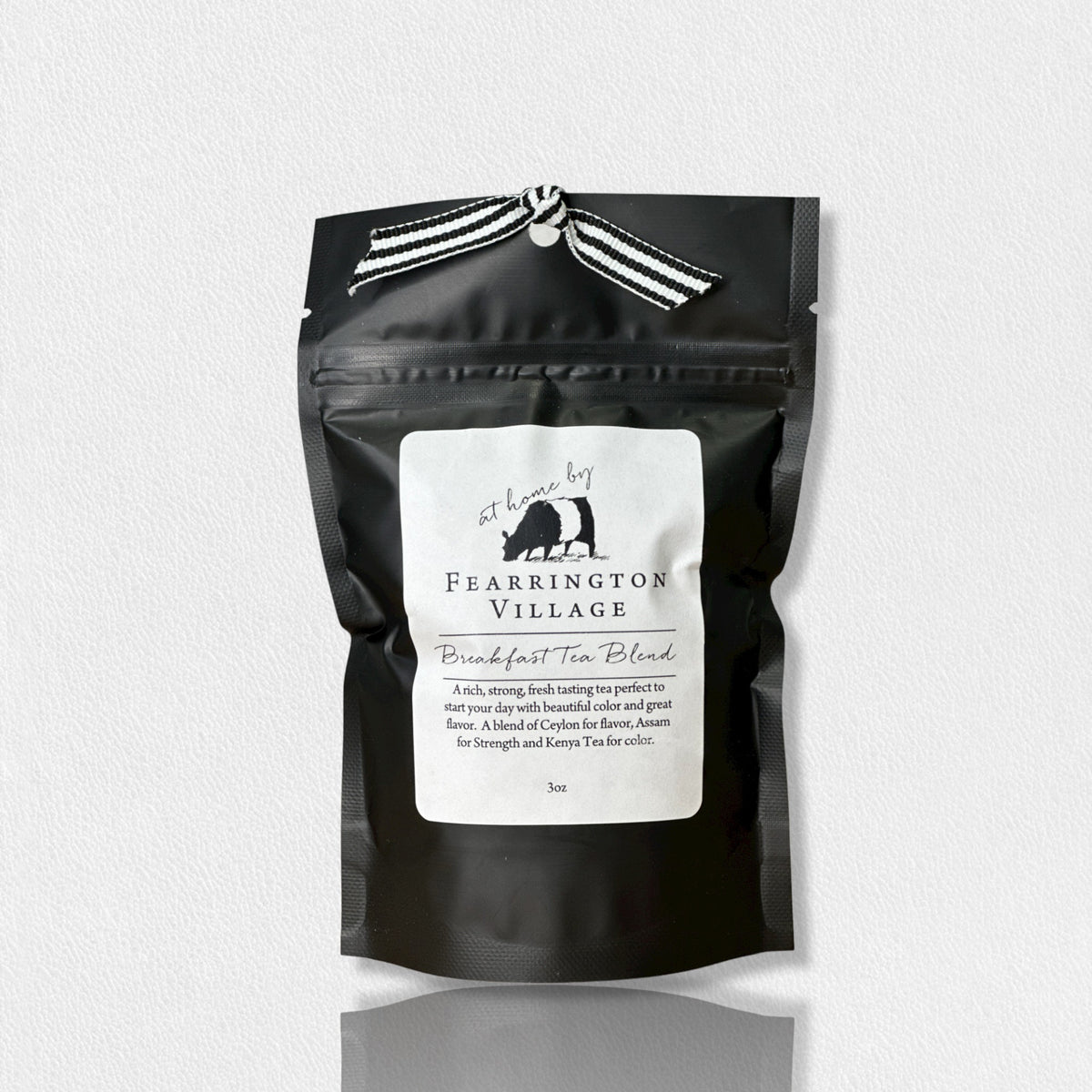 A black resealable bag of the Fearrington Signature Collection - Fearrington Signature Breakfast Tea Blend. The white label prominently displays the Fearrington House brand and describes it as &quot;An elegant blend of Ceylon and Assam with a hint of Keemun for strength.&quot; This loose leaf tea is adorned with a striped ribbon on top, offering a rich, strong tea experience.