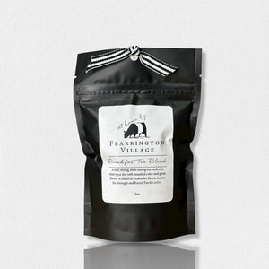 A black resealable bag of the Fearrington Signature Collection - Fearrington Signature Breakfast Tea Blend. The white label prominently displays the Fearrington House brand and describes it as "An elegant blend of Ceylon and Assam with a hint of Keemun for strength." This loose leaf tea is adorned with a striped ribbon on top, offering a rich, strong tea experience.