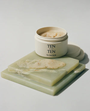 A jar of TENOVERTEN - THE SOLE SCRUB by TEN OVER TEN is displayed on a green marble surface. The jar lid is off, revealing the scrub inside. Some of the scrub is spread out on a slab next to the jar, showcasing its creamy, coarse texture infused with orange peel oil for a revitalizing exfoliation and smoother skin texture.
