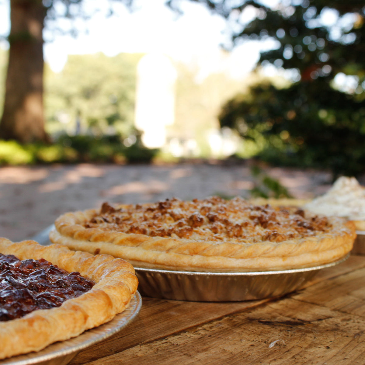 A fruit pie and a crumb-topped Thanksgiving Pies - Pickup November 26, 2026 on a wooden table outdoors.