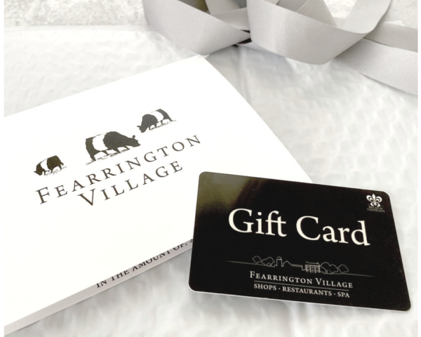 A Fearrington Village gift card and matching envelope are displayed on a white textured surface with a silver ribbon in the background. The gift card and envelope both feature the Fearrington Village logo.