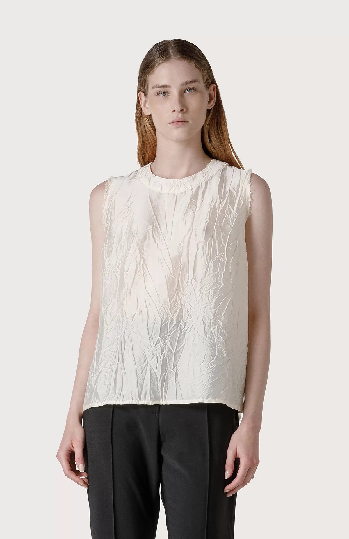 A person with long, straight hair wears a sleeveless, textured SLEEVELESS TOP by SEVENTY and black pants. They are posing against a plain, light grey background. The top features raw-cut armholes and a crinkled appearance, adding depth and visual interest to the outfit.