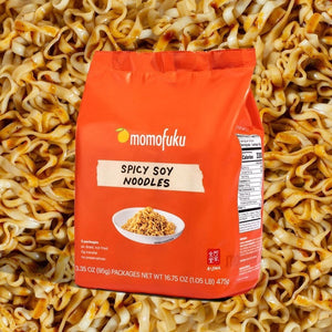 A vibrant orange package of MOMOFUKU Spicy Soy Noodles highlights savory and spicy flavors. The front features the MOMOFUKU brand logo, a small white bowl of noodles, and various product details. It includes five individual packs of air-dried noodles, each weighing 95 grams, with a total weight of 475 grams.