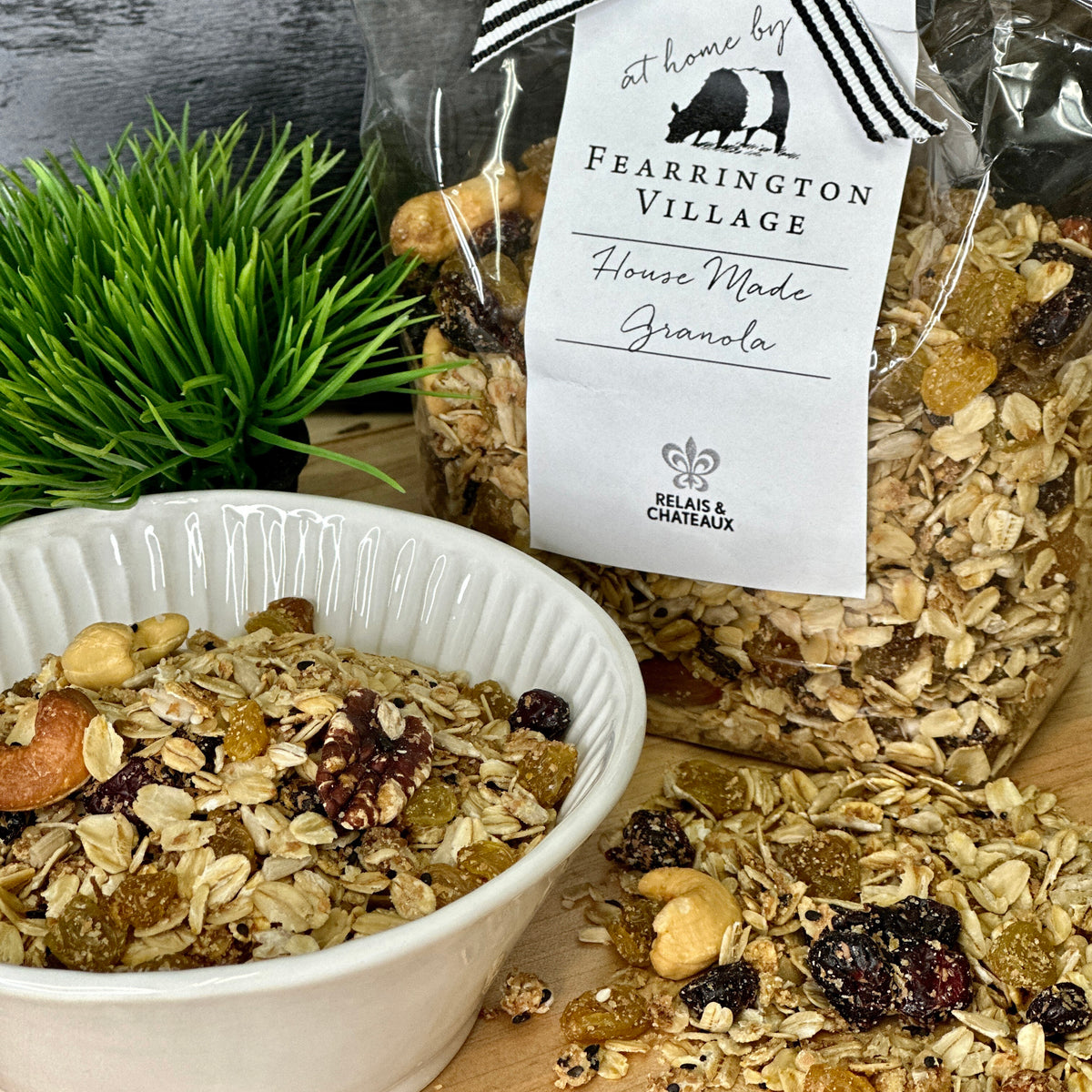 A clear plastic container with a silver lid, labeled "Fearrington Signature Collection - Fearrington Housemade Granola." The label lists ingredients such as oatmeal, sesame seeds, and various nuts. Brimming with gourmet granola, the container is set against a plain white background.