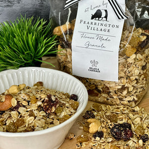 A clear plastic container with a silver lid, labeled "Fearrington Signature Collection - Fearrington Housemade Granola." The label lists ingredients such as oatmeal, sesame seeds, and various nuts. Brimming with gourmet granola, the container is set against a plain white background.