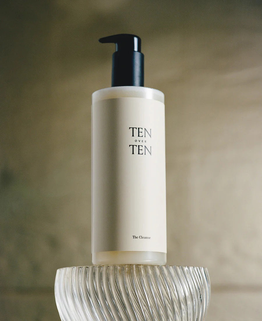 A beige bottle with a black pump top, labeled &quot;TENOVERTEN - THE CLEANSE HAND WASH&quot; from the brand TEN OVER TEN, sits atop a textured, translucent base. This hand wash features botanical extracts for skin hydration. The background is softly blurred, drawing attention to the elegantly designed bottle.