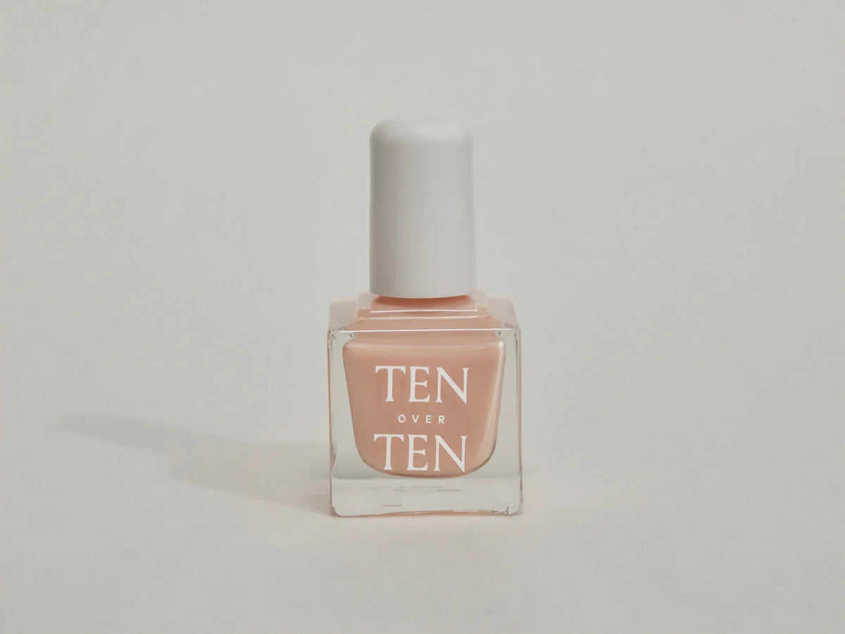 A glass bottle of TENOVERTEN - NAIL POLISH from the brand TEN OVER TEN, featuring a white cap and a dusty pink nail color. The front label displays "TEN OVER TEN" in white uppercase letters. This vegan, eight-free formula ensures both beauty and peace of mind. The background is plain and light-colored.