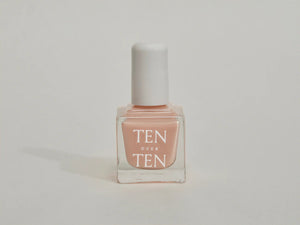 A glass bottle of TENOVERTEN - NAIL POLISH from the brand TEN OVER TEN, featuring a white cap and a dusty pink nail color. The front label displays "TEN OVER TEN" in white uppercase letters. This vegan, eight-free formula ensures both beauty and peace of mind. The background is plain and light-colored.