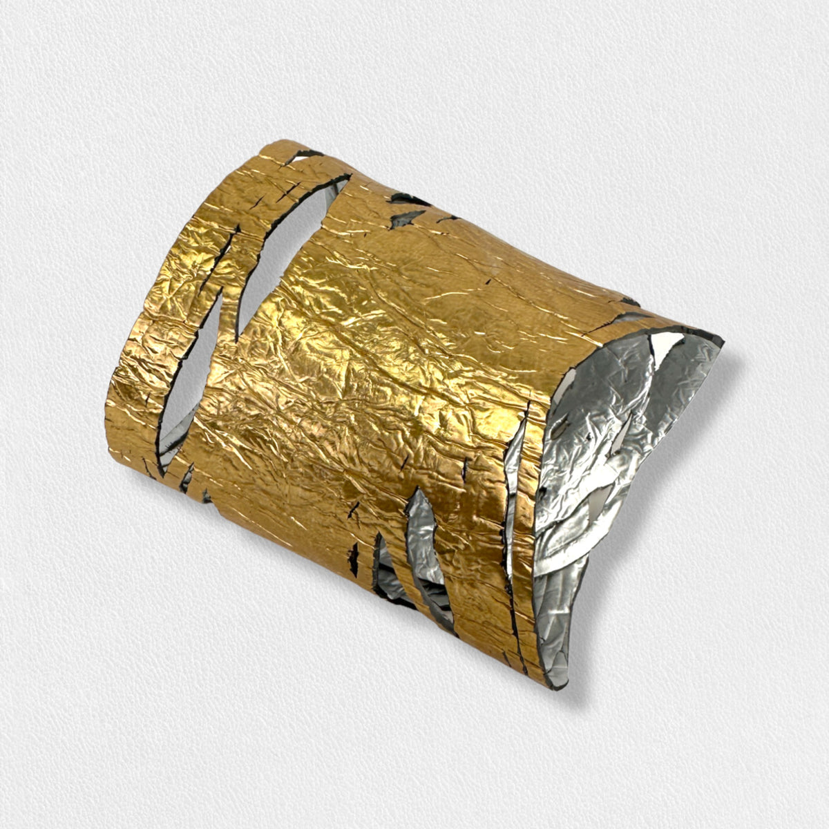 A person wearing the ILLUSIONISM BRACELET IN GOLD AND SILVER (REVERSIBLE) from SI SABATO ISABEL on their wrist. The handmade jewellery, crafted by an Argentinian artist, features irregular cut-out shapes and an intricate, hammered surface. The person is dressed in a faded blue denim shirt with white buttons.