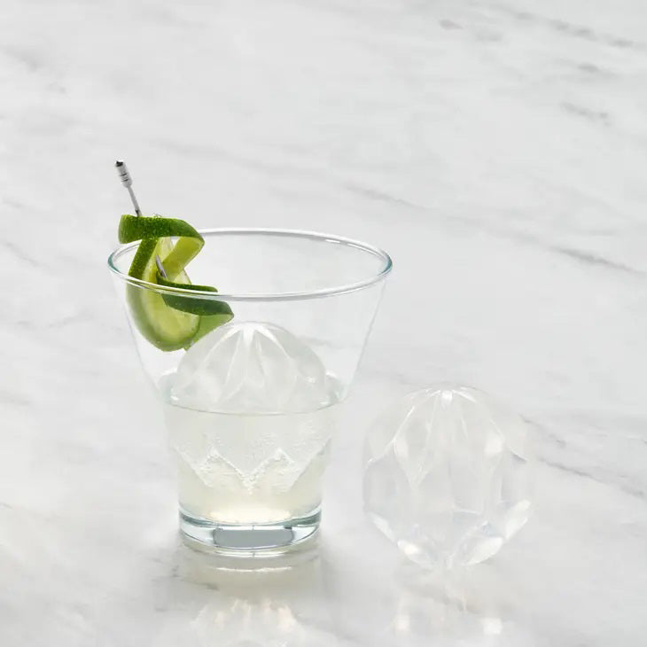 A black, hexagonal W&amp;P - Ripple Cocktail Ice Mold with its top half removed, revealing a clear ice cube inside shaped like a decorative sphere with petal-like engravings. Another identical ice cube is shown next to the mold, perfect for chilling drinks with its intricate sculptural wave pattern from W&amp;P Design.
