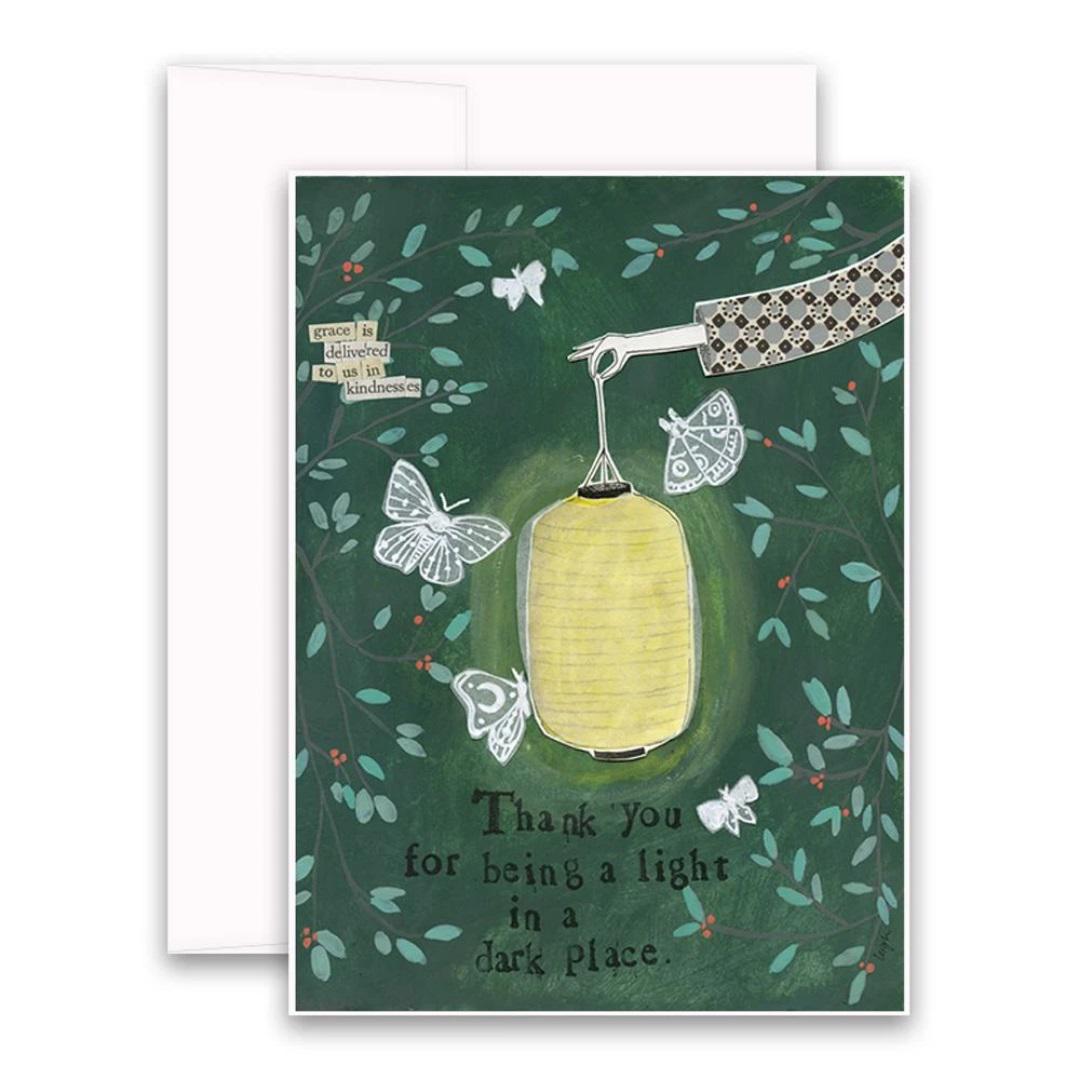 The CURLY GIRL DESIGNS - LIGHT IN THE DARK CARD by CURLY GIRL DESIGN, INC showcases a beautiful collage art illustration of a hand holding a glowing lantern surrounded by butterflies. The card is adorned with the heartfelt message, "Thank you for being a light in a dark place." Additionally, above the hand, there is a small sign that reads, "Grace is delivered in its kindness.