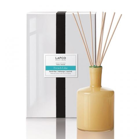 A beige LAFCO - French Lilac, Poolhouse Diffuser with thin wooden sticks is placed in front of a white box showcasing a black vertical stripe. The labeled &quot;LAFCO New York - French Lilac&quot; box emphasizes the heliotrope and hyacinth top notes that contribute to the enchanting French lilac fragrance.
