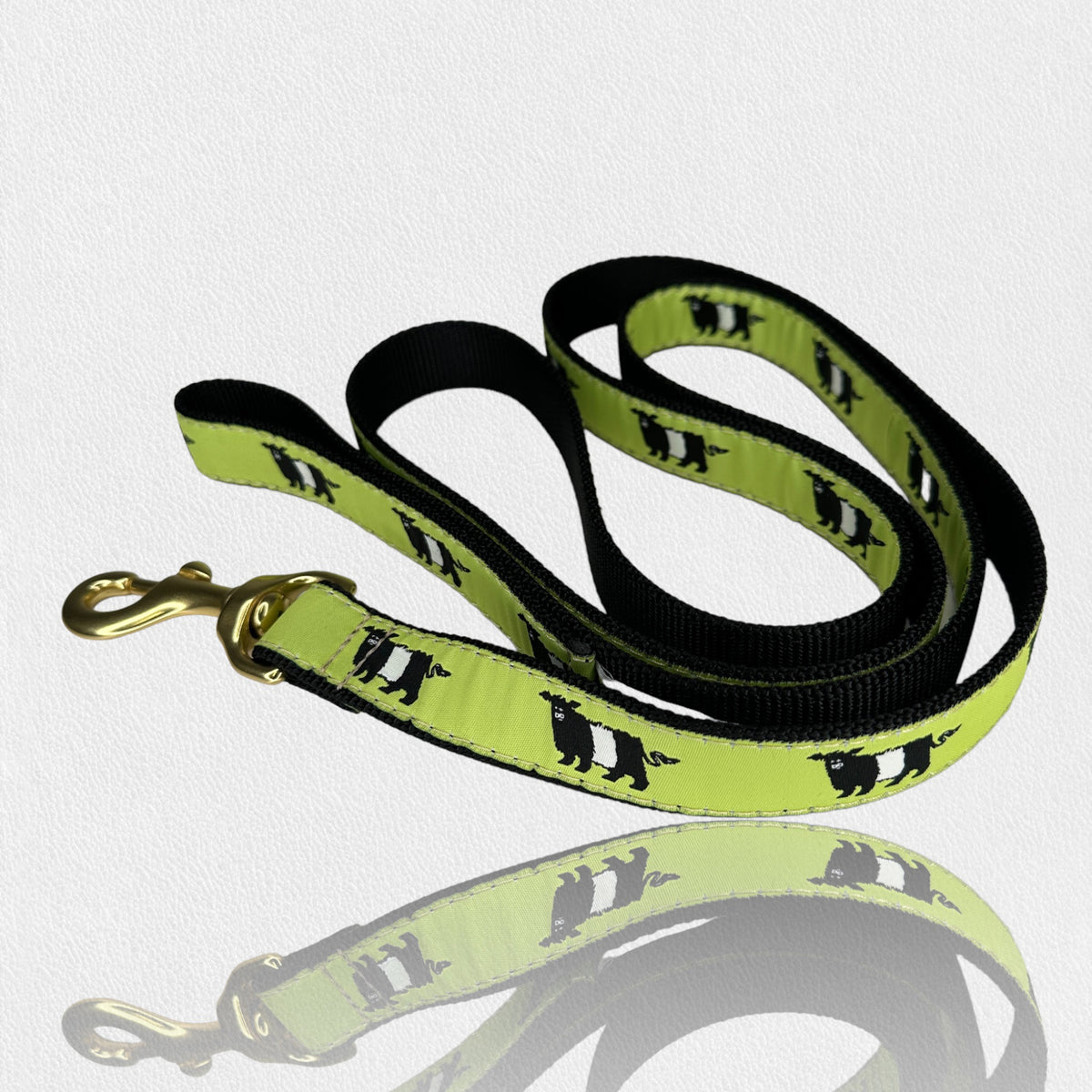 A FEARRINGTON SIGNATURE COLLECTION - BESPOKE BELTIE DOG LEASH from UP COUNTRY is displayed on a white surface. This coiled, black and green dog leash features a gold-tone clasp and a repeating pattern of black sheep silhouettes on the green sections. Ideal for dog lovers, it pairs perfectly with a matching collar for a coordinated look. The leash&#39;s shadow is reflected below it.