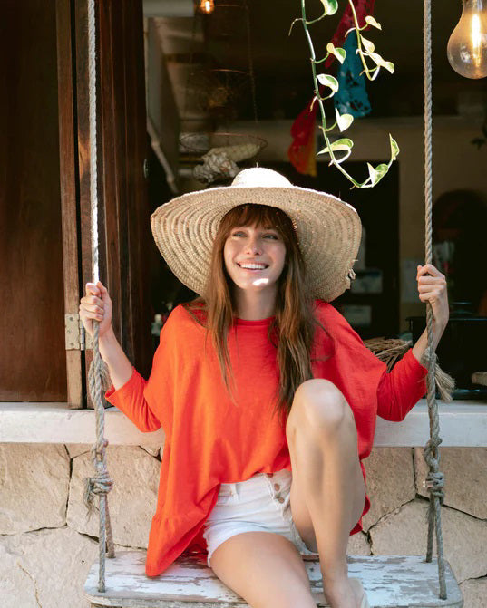 Wearing the MERSEA MOROCCAN RUSTIC STRAW HAT, a woman in an orange top and white shorts smiles on a swing, surrounded by plants and hanging lights, enjoying a bright sunny day.