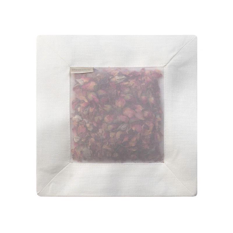 The ROSE SQUARE SACHET IVORY LINEN by E WIGHTMAN &amp; CO features a white fabric cover with a mesh window that displays dried lavender buds and scented rose petals inside. A small beige tag is attached on the top left side of the sachet.