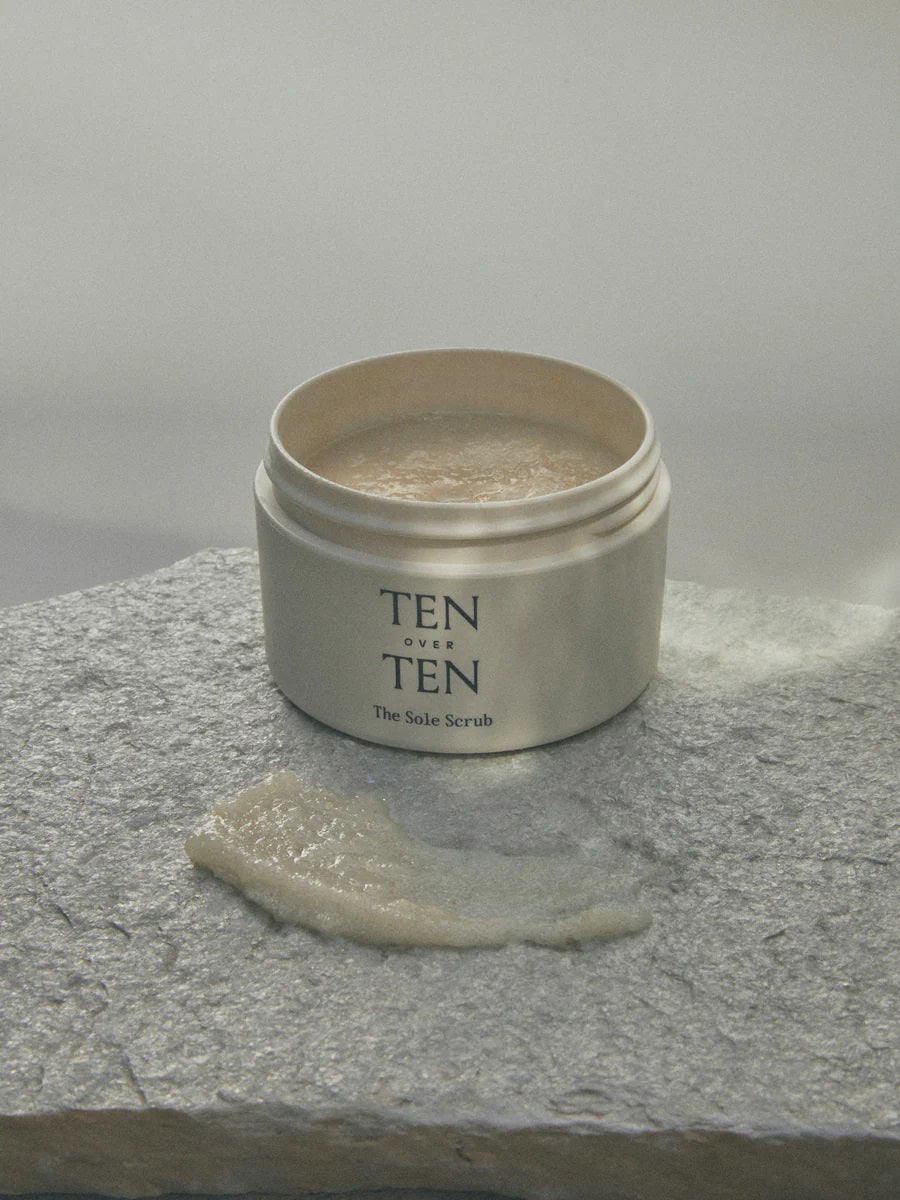 A jar of TENOVERTEN - THE SOLE SCRUB by TEN OVER TEN is displayed on a green marble surface. The jar lid is off, revealing the scrub inside. Some of the scrub is spread out on a slab next to the jar, showcasing its creamy, coarse texture infused with orange peel oil for a revitalizing exfoliation and smoother skin texture.