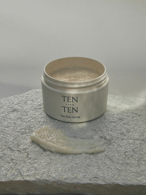 A jar of TENOVERTEN - THE SOLE SCRUB by TEN OVER TEN is displayed on a green marble surface. The jar lid is off, revealing the scrub inside. Some of the scrub is spread out on a slab next to the jar, showcasing its creamy, coarse texture infused with orange peel oil for a revitalizing exfoliation and smoother skin texture.