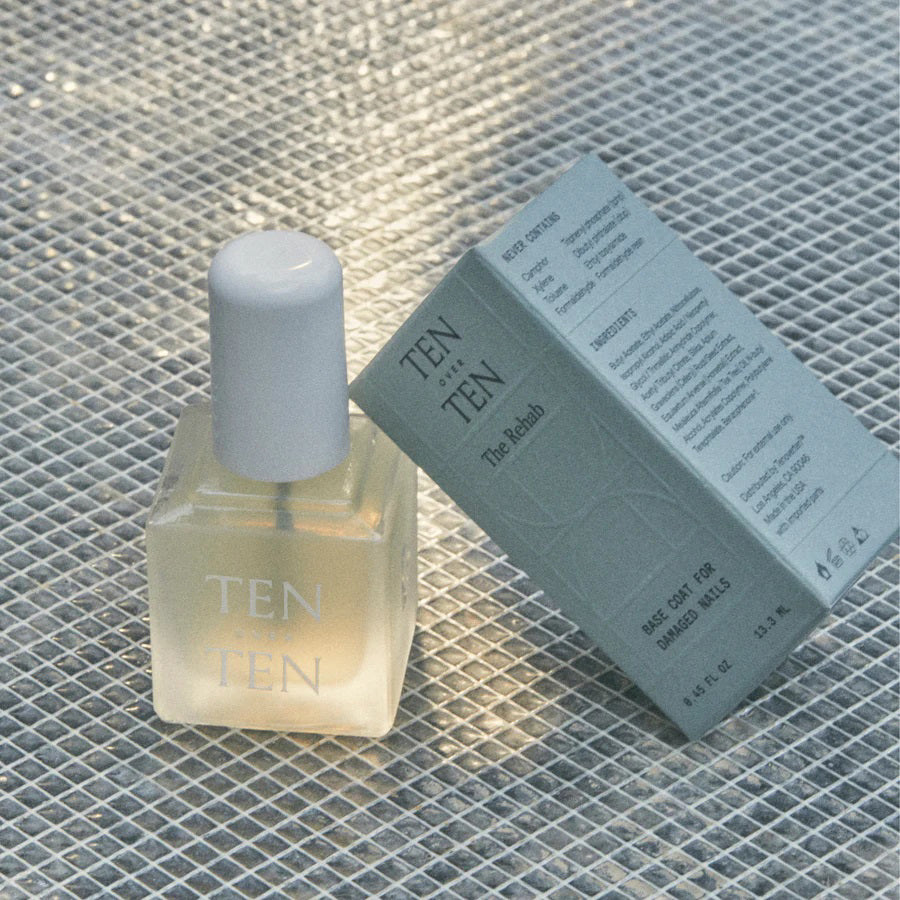 A bottle of TENOVERTEN - THE REHAB BASE COAT with a white cap is placed next to a light green box featuring the brand name 'TEN OVER TEN' and product details, perfect for those seeking to maintain nail health. The background is a textured, reflective surface.