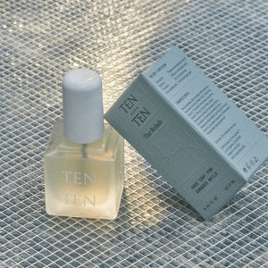 A bottle of TENOVERTEN - THE REHAB BASE COAT with a white cap is placed next to a light green box featuring the brand name 'TEN OVER TEN' and product details, perfect for those seeking to maintain nail health. The background is a textured, reflective surface.