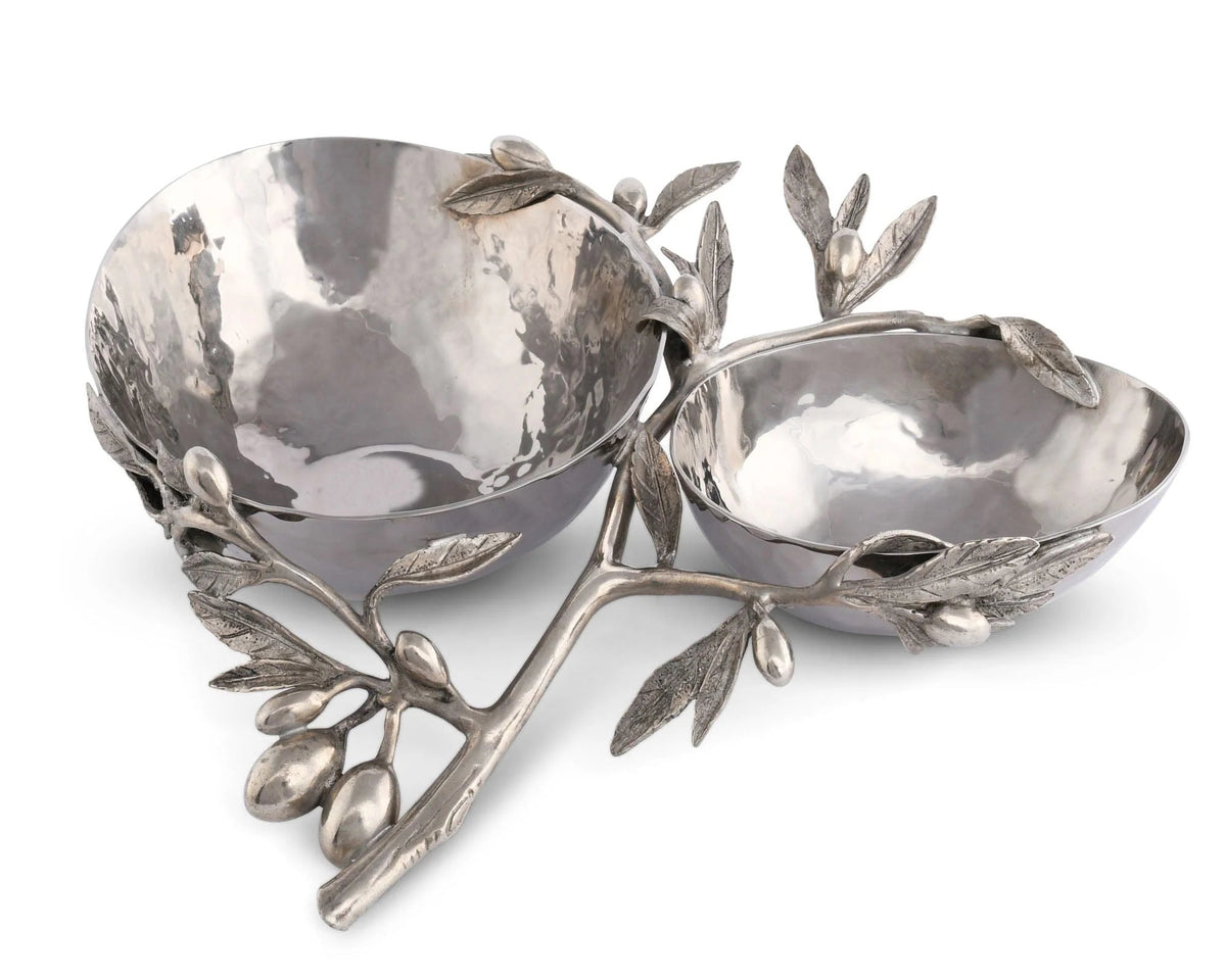 The Olive Double Serving Bowl from Vagabond House features two gleaming silver metal bowls linked by a beautifully intricate branch design made of pure pewter. Delicate leaves and tiny olives artistically entwine, creating an elegant stand for this stunning piece. Hand washing is recommended to preserve its beauty.