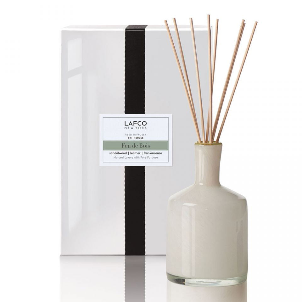 A white ceramic diffuser bottle with reed sticks is displayed in front of a minimalistic, white rectangular box. The box features a vertically centered black ribbon and a label that reads, "LAFCO New York, Feu de Bois, Sandalwood, Leather, Virginia Cedarwood." This LAFCO Ski House Feu de Bois Diffuser is perfect for creating a warm ambiance.