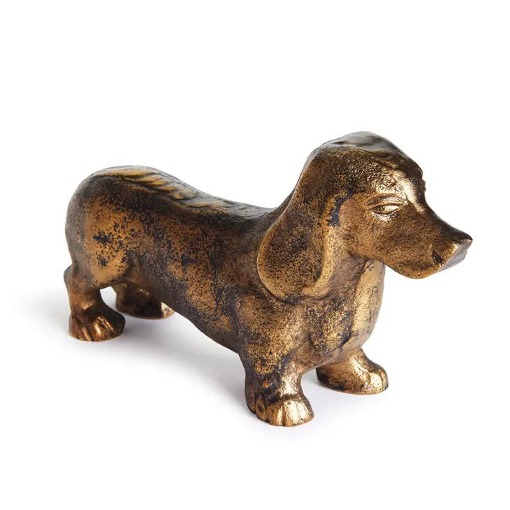 The NAPA HOME AND GARDEN - MOXIE BRASS SCULPTURE is a compact cast aluminum dachshund with a textured finish, capturing its long body, floppy ears, and delicate features. This blend of realism with artistic style makes it ideal as charming bookshelf decor.