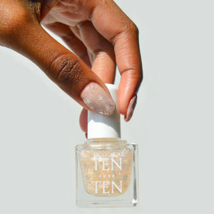 A glass bottle of TENOVERTEN - NAIL POLISH from the brand TEN OVER TEN, featuring a white cap and a dusty pink nail color. The front label displays "TEN OVER TEN" in white uppercase letters. This vegan, eight-free formula ensures both beauty and peace of mind. The background is plain and light-colored.