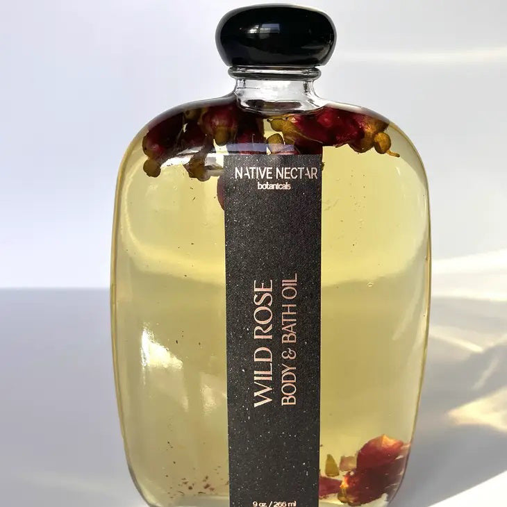 A glass bottle filled with luxurious, soothing oil. The clear liquid inside showcases petals and botanical extracts. The bottle features a black cap and a dark label reading "NATIVE NECTAR" and "WILD ROSE BODY OIL." The label specifies the quantity as 9 fl oz / 220 ml.