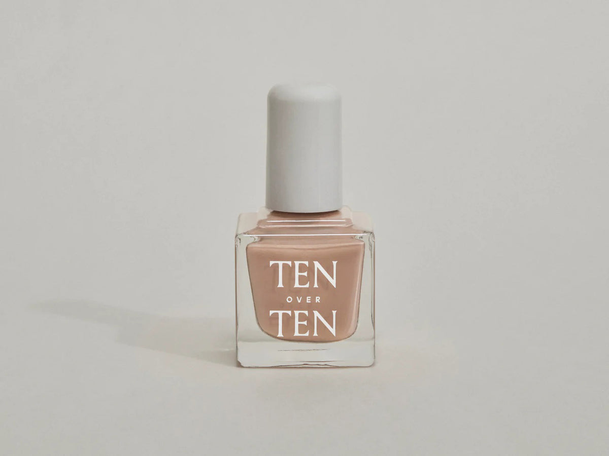 A glass bottle of TENOVERTEN - NAIL POLISH from the brand TEN OVER TEN, featuring a white cap and a dusty pink nail color. The front label displays "TEN OVER TEN" in white uppercase letters. This vegan, eight-free formula ensures both beauty and peace of mind. The background is plain and light-colored.