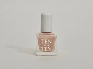 A glass bottle of TENOVERTEN - NAIL POLISH from the brand TEN OVER TEN, featuring a white cap and a dusty pink nail color. The front label displays "TEN OVER TEN" in white uppercase letters. This vegan, eight-free formula ensures both beauty and peace of mind. The background is plain and light-colored.