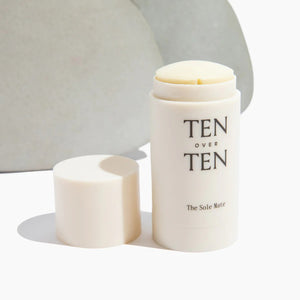 An open white cylindrical container displaying the brand "TEN OVER TEN" and product name "TENOVERTEN - THE SOLE MATE." The lid rests beside the container, revealing a solid cream stick enriched with botanical oils. The minimalist, light-colored background accentuates how effectively you can moisturize feet.